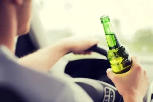 A driver with one hand on the steering wheel and a beer bottle in the other