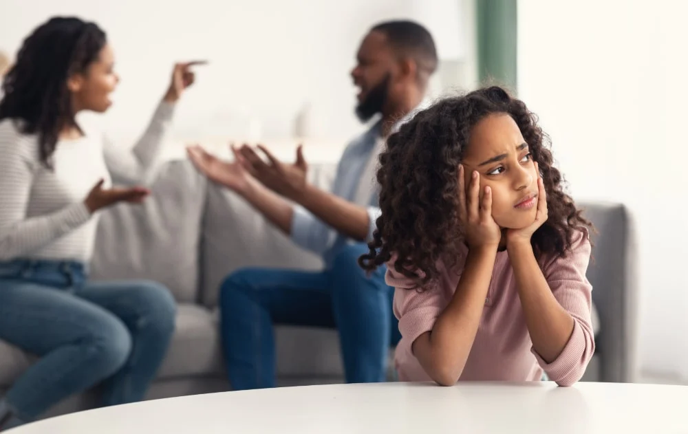 A girl holding her head in her hands while her parents argue behind her