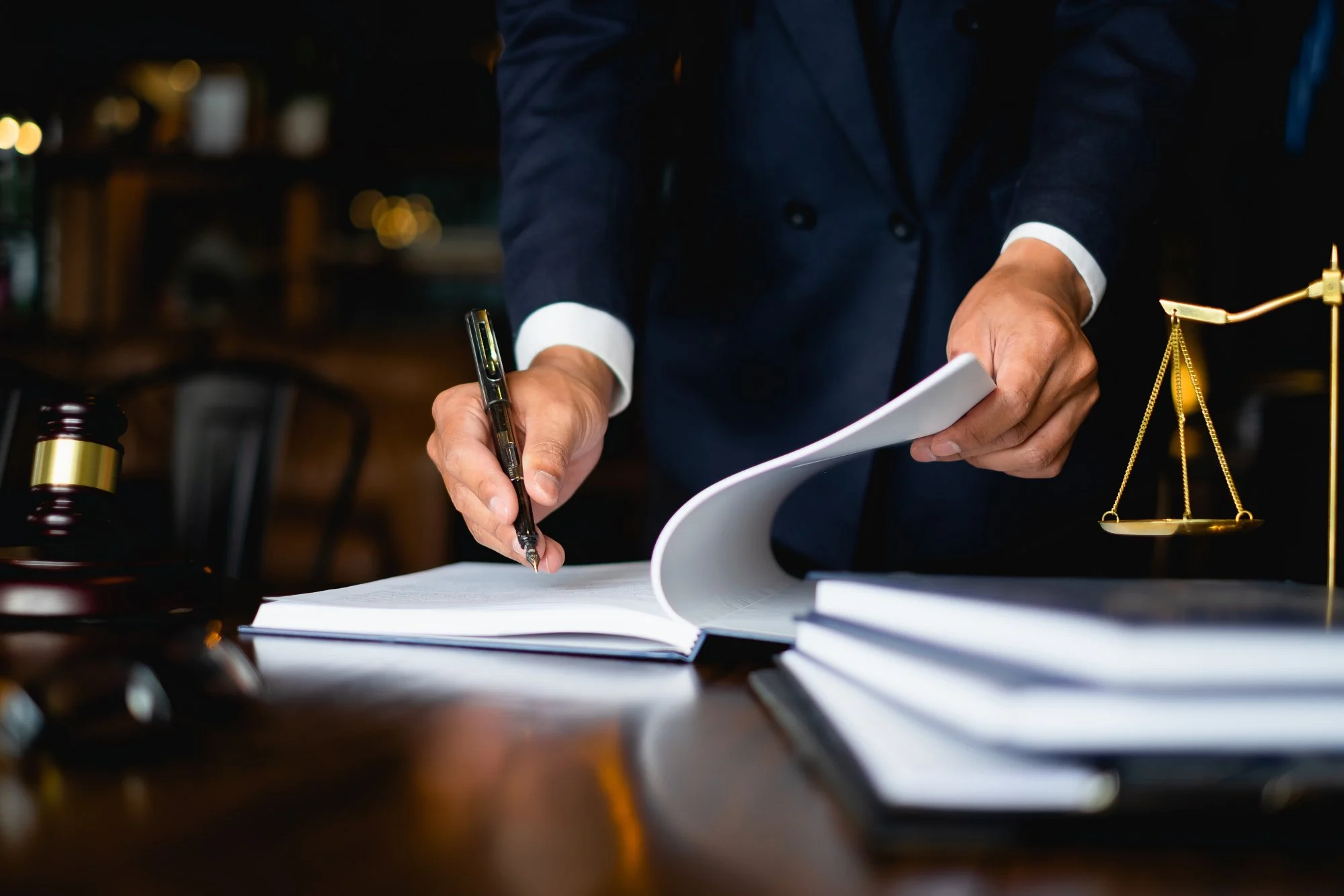 A lawyer in a suit signing papers at a desk beside a gavel and the scales of justice