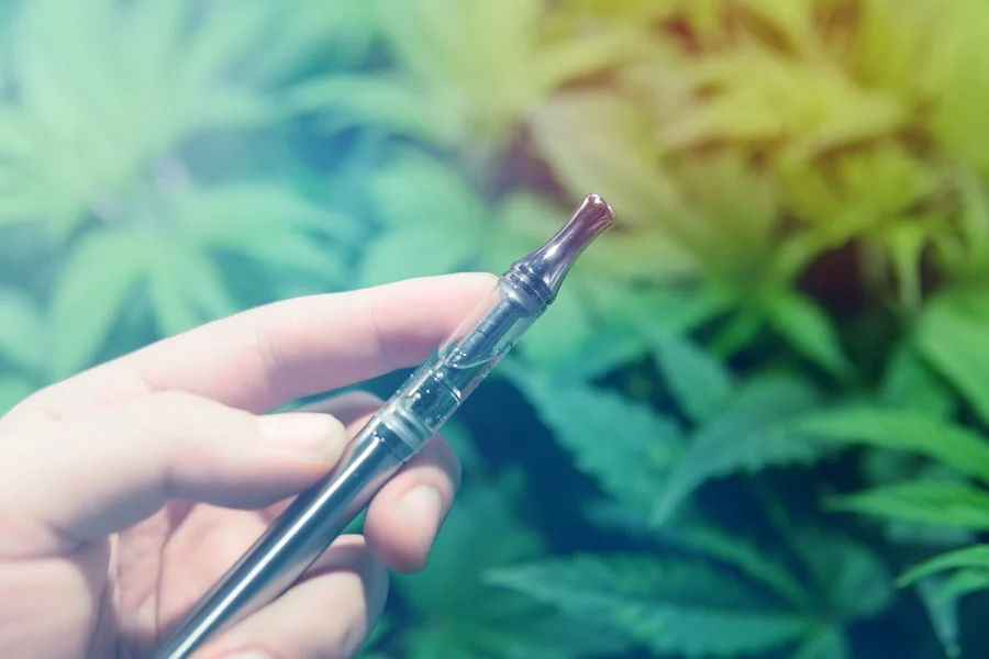 A hand holding a vape pen in front of cannabis leaves