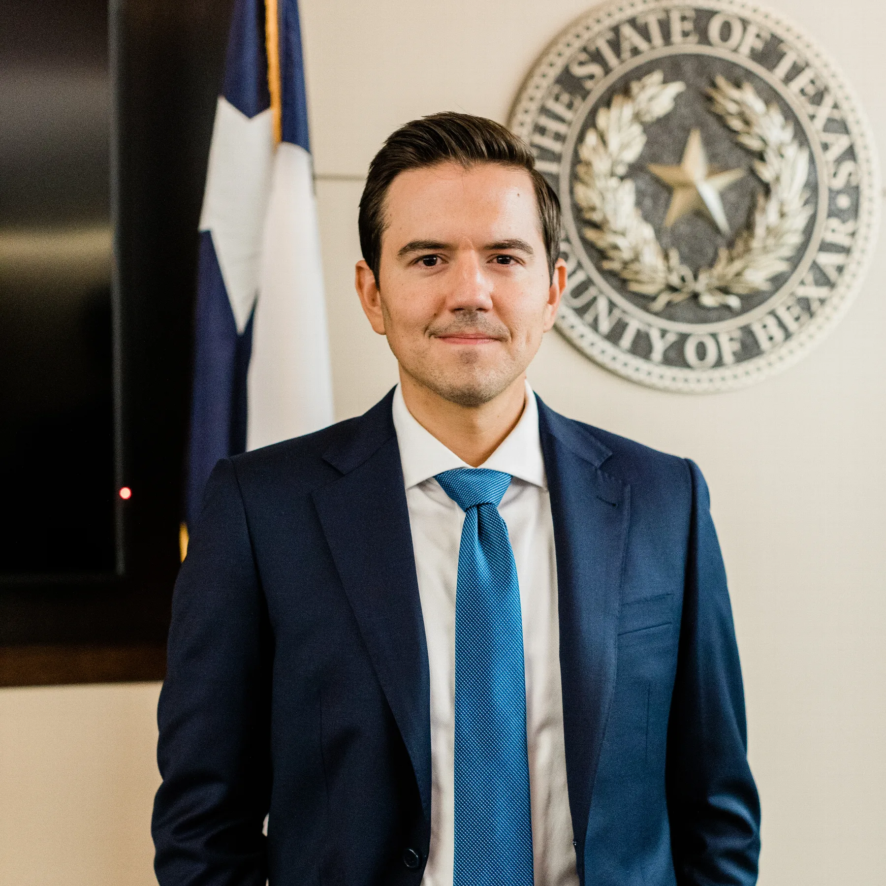 Austin Hagee standing before the Texas flag and the Bexar County seal